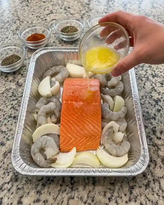 Raw salmon fillet with shrimp and onion wedges in a foil pan, melted butter being poured on top, seasoning bowls in background