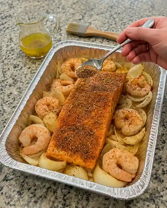 Hand spooning paprika seasoning blend over a salmon fillet surrounded by shrimp in a foil baking pan