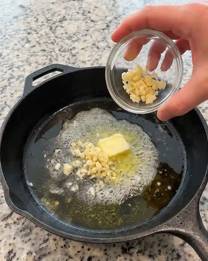 Minced garlic being added to melted butter and olive oil in a cast iron skillet